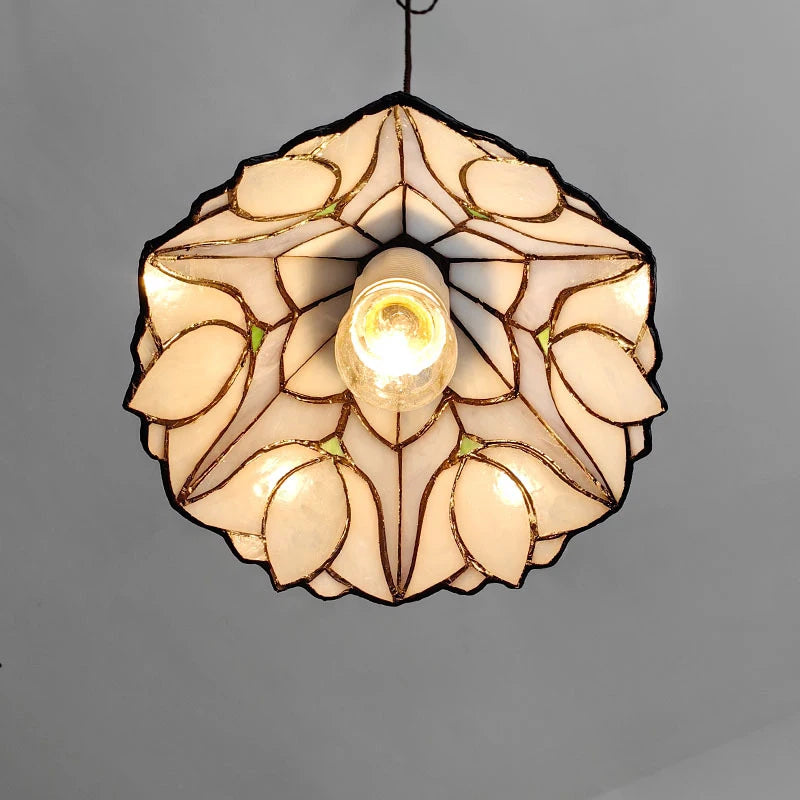 A circular pendant light shade with a stained-glass tulip pattern, featuring warm cream-colored glass panels outlined in dark metal with small green accents, illuminated from the center against a plain gray background.