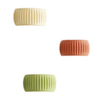 Three ribbed cylindrical wall sconces in cream, terracotta, and light green, shown mounted on a white wall with warm light glowing from each.