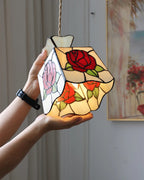 A stained glass pendant light shaped like a teardrop, featuring red and pink roses with green leaves, held by two hands against an indoor background.