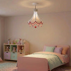 A whimsical children's pendant light hanging above a bed in a pastel bedroom. The light fixture has a white fabric shade with red scalloped trim and dangling pink and red pom-poms, suspended by a black cord.