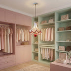 A decorative pendant chandelier with a cream-colored bell-shaped shade, red scalloped trim, and pink pom-pom accents hanging in a pastel pink and mint green walk-in closet.