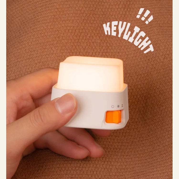 A hand holding a small white key light device that glows with warm white light, featuring an orange accent switch, set against a brown textured background with 'KEYLIGHT !!!' text overlay.
