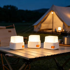 Three small white camping lanterns with orange switches are lit and placed on a folding table outdoors at dusk, with a glowing tent and campsite setting in the background.