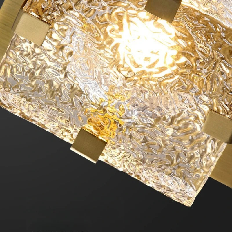 A close-up of a modern square pendant light with a textured, hammered clear glass shade and brushed gold metal brackets, illuminated against a dark background.