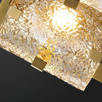 A close-up of a modern square pendant light with a textured, hammered clear glass shade and brushed gold metal brackets, illuminated against a dark background.