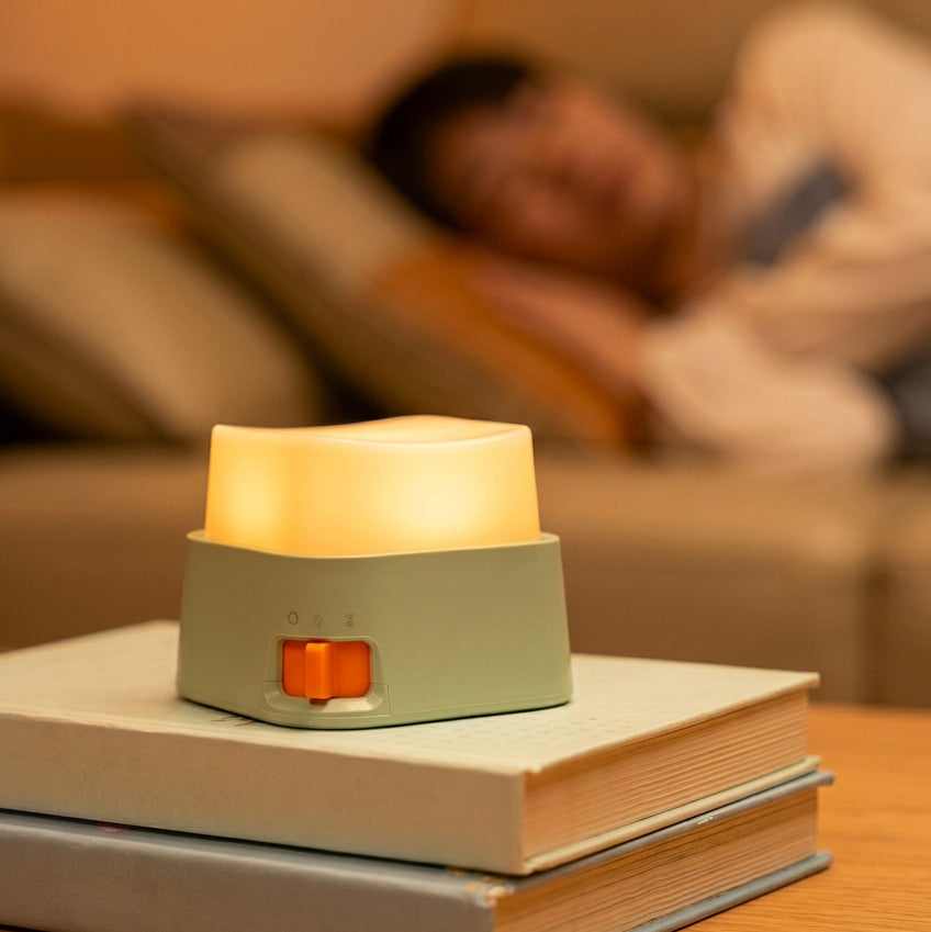 A small green and white night light with an orange switch sits on top of a light blue book in the foreground. The background is a softly blurred living room scene with a person lying on a couch, and large text reads '15 minutes Delay lights out' with a smaller explanatory blurb about automatically turning off the lights in 15 minutes.