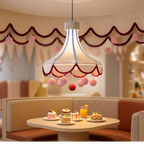 A whimsical pendant light with a translucent white bell-shaped shade, red scalloped trim, and dangling pink and red pom-poms, hanging above a cafe table with desserts and drinks in a cozy interior setting.