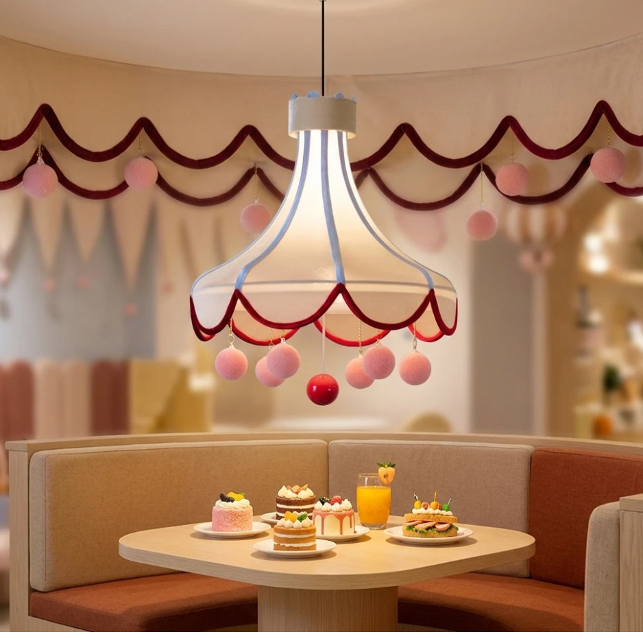 A whimsical pendant light with a translucent white bell-shaped shade, red scalloped trim, and dangling pink and red pom-poms, hanging above a cafe table with desserts and drinks in a cozy interior setting.