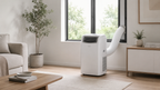 A white portable window air conditioner with a flexible white hose extending out a tall window in a bright, modern living room.